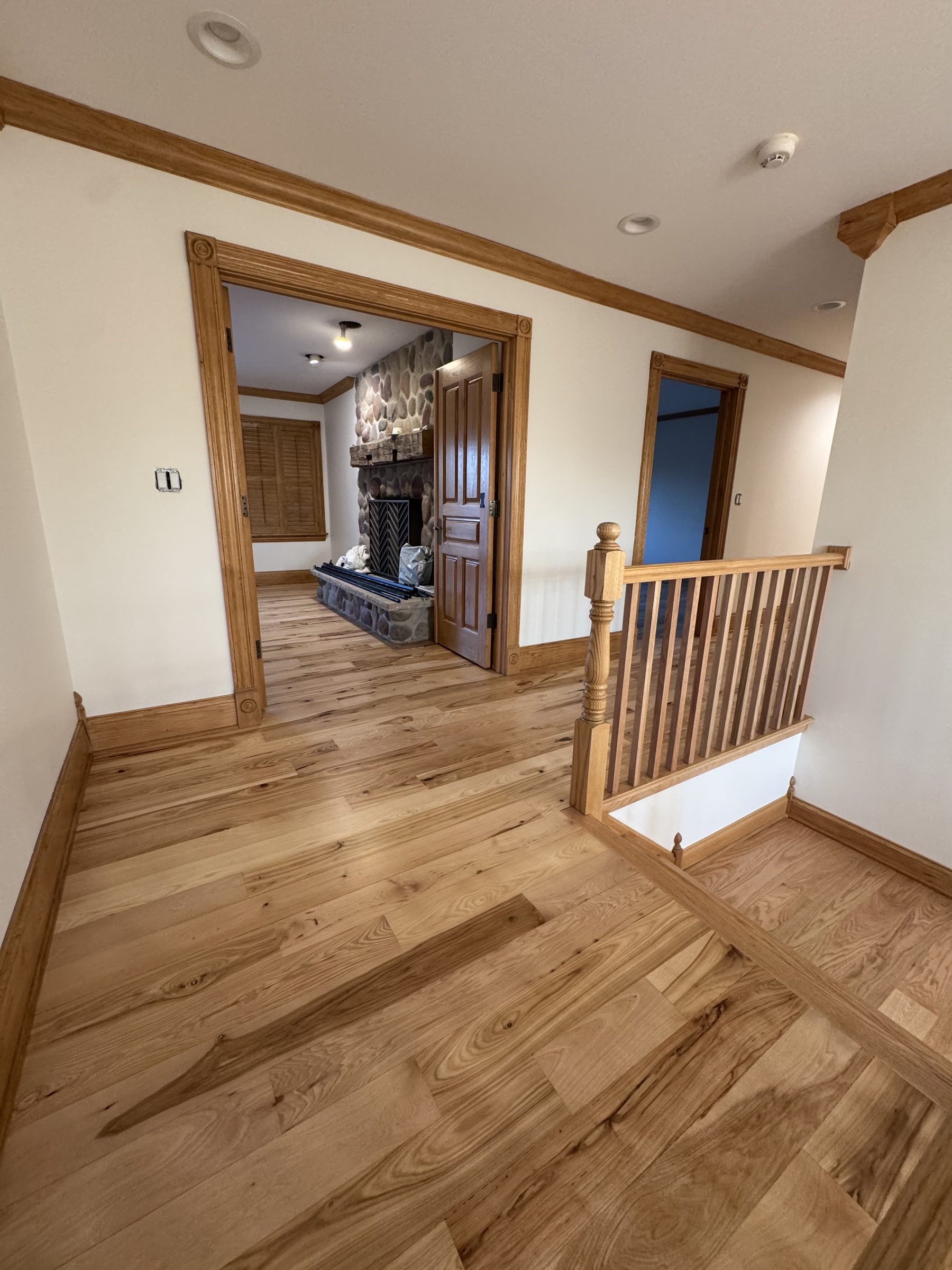 Wide-plank natural hickory floor running through an upstairs hallway and into a room with a stone fireplace, with a matching oak railing